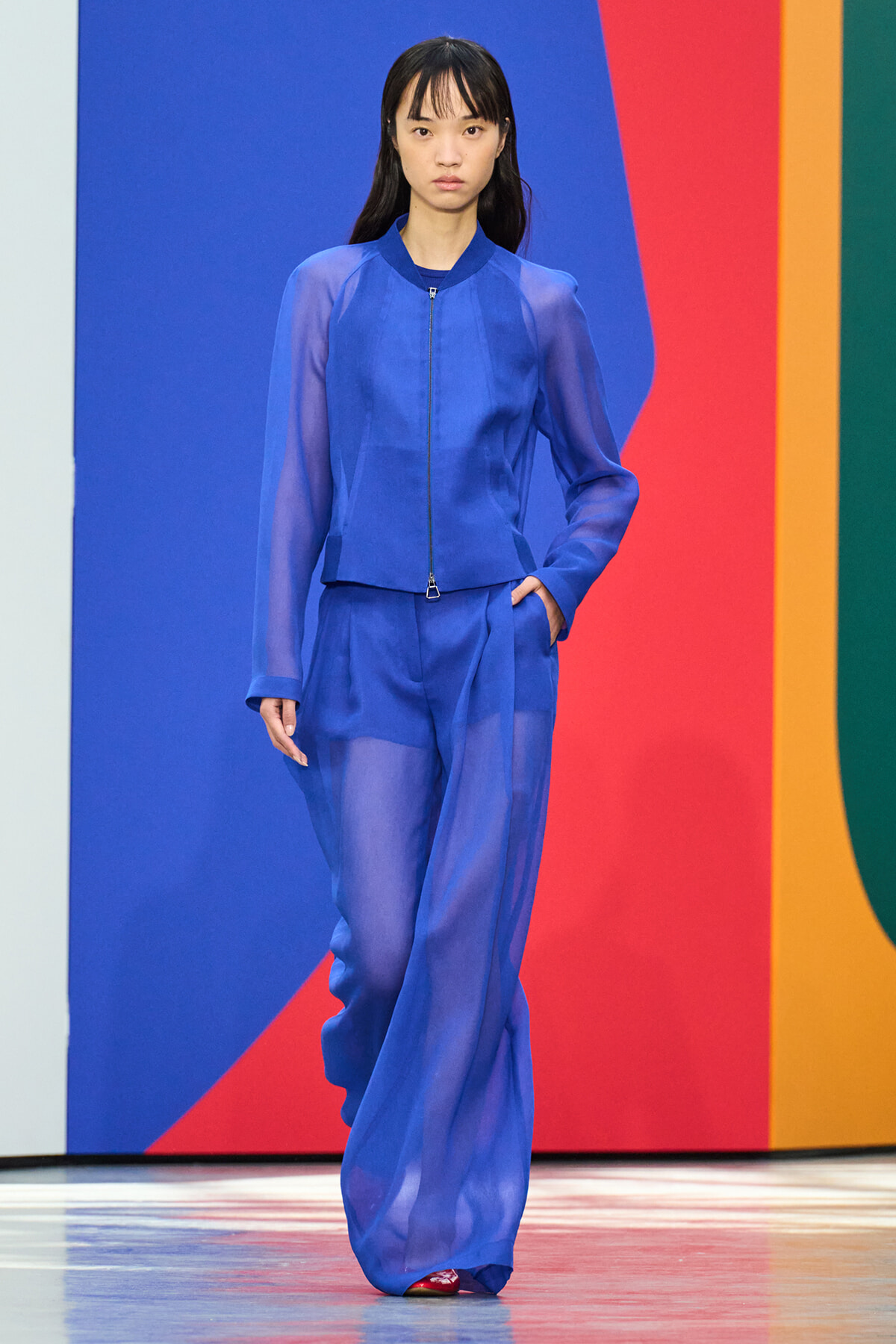 Model walks a runway in a cobalt blue sheer jacket and wide-leg pants against a colorful geometric backdrop.