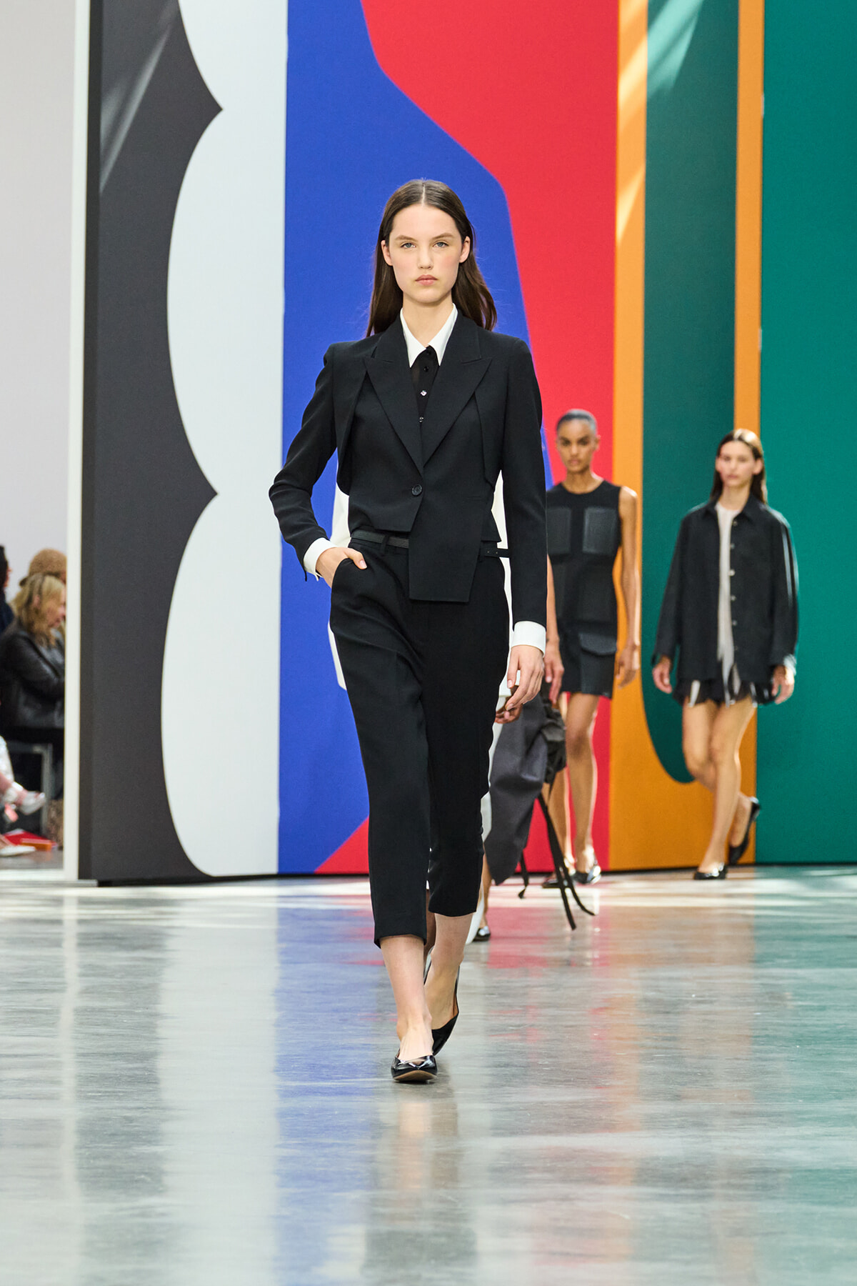 Model in a tailored black suit walks down a glossy runway with a colorful abstract backdrop behind her.