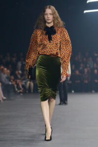 Model walking a runway in an orange polka-dot blouse with a black bow, paired with a green velvet pencil skirt and black heels, carrying a black handbag.