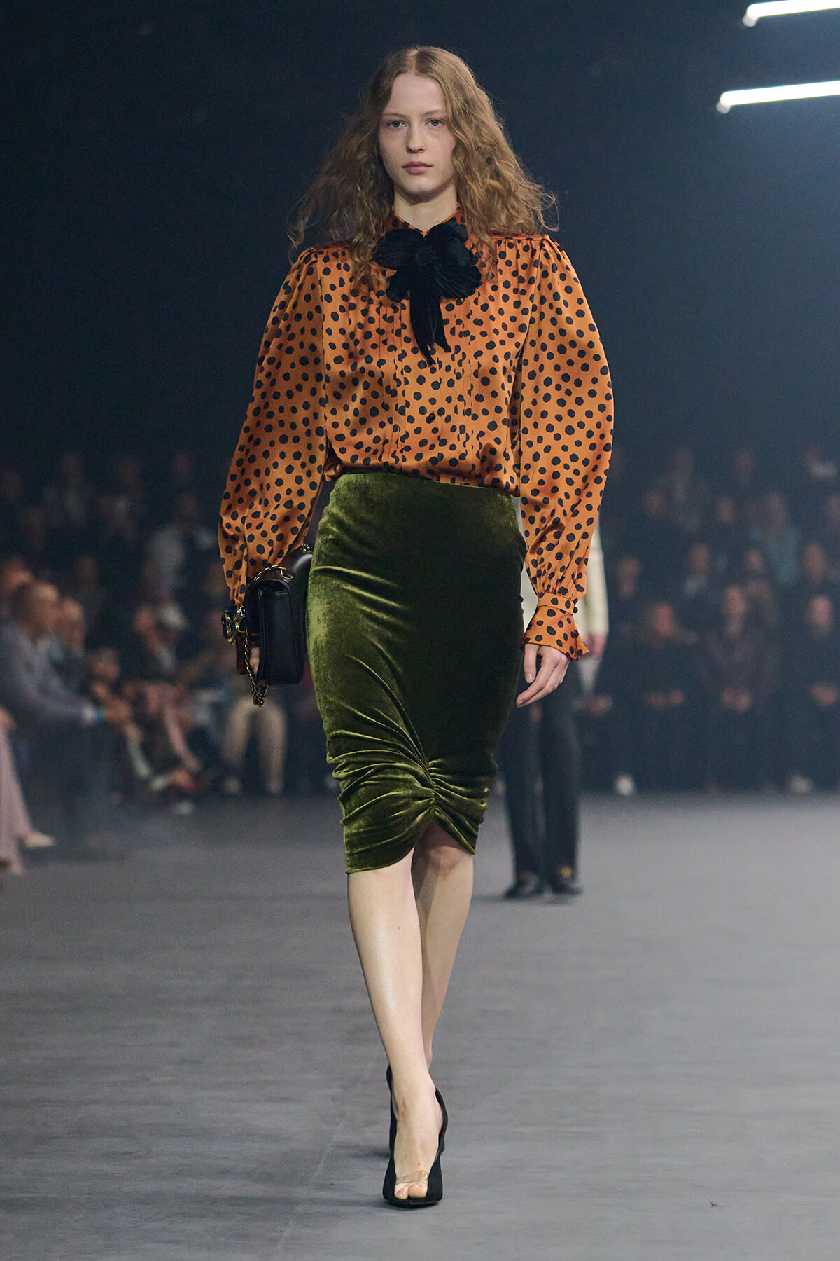 Model walking a runway in an orange polka-dot blouse with a black bow, paired with a green velvet pencil skirt and black heels, carrying a black handbag.