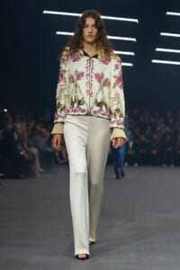 Female model on a runway wearing a sequined white floral jacket with pink flowers and cream fringe cuffs, paired with wide-leg ivory pants.