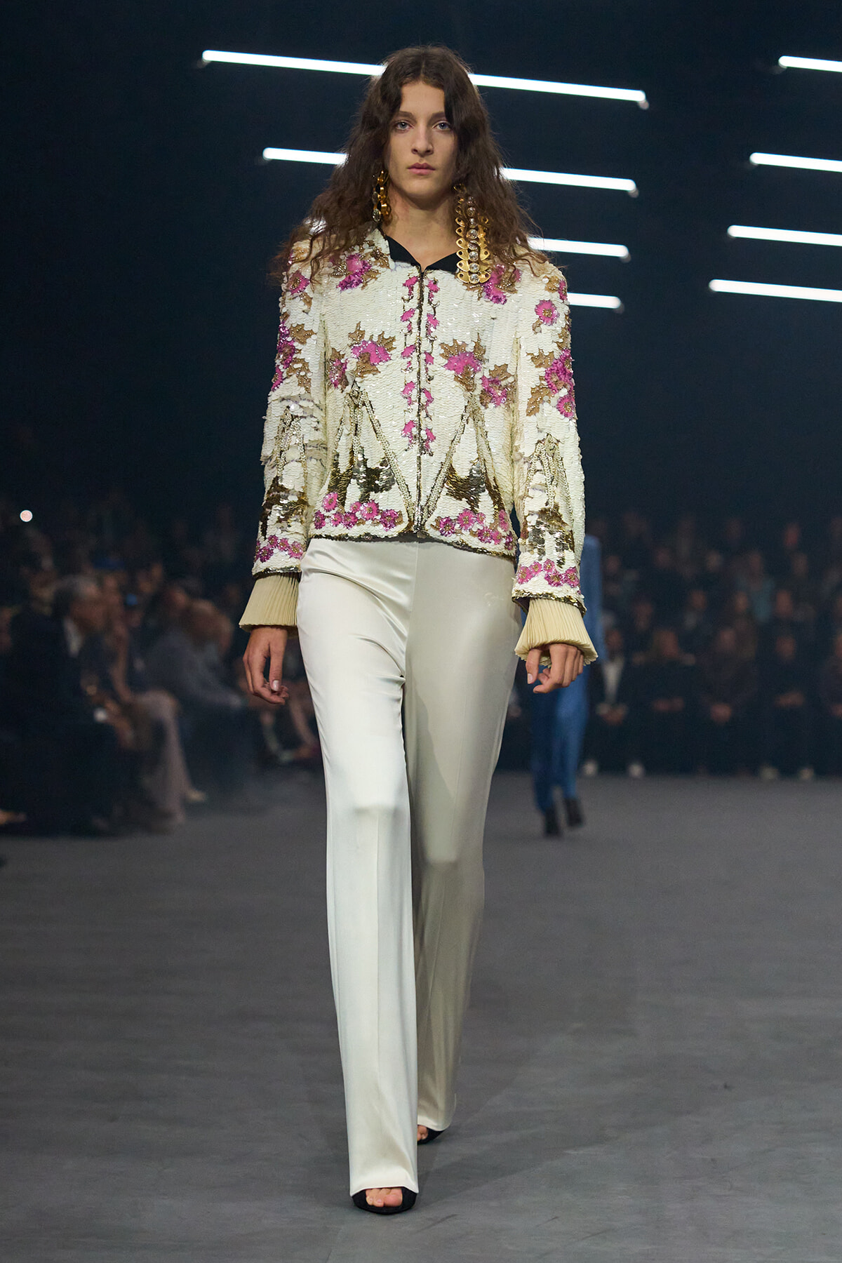 Female model on a runway wearing a sequined white floral jacket with pink flowers and cream fringe cuffs, paired with wide-leg ivory pants.