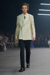 Male model walks the runway in a white double-breasted dotted blazer, black trousers, and black loafers with gold hardware, holding a clutch.