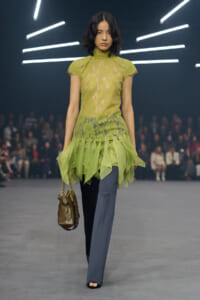Model walking runway in lime lace high-neck top over sheer skirted hem, blue-gray wide-leg pants, brown handbag with chain.