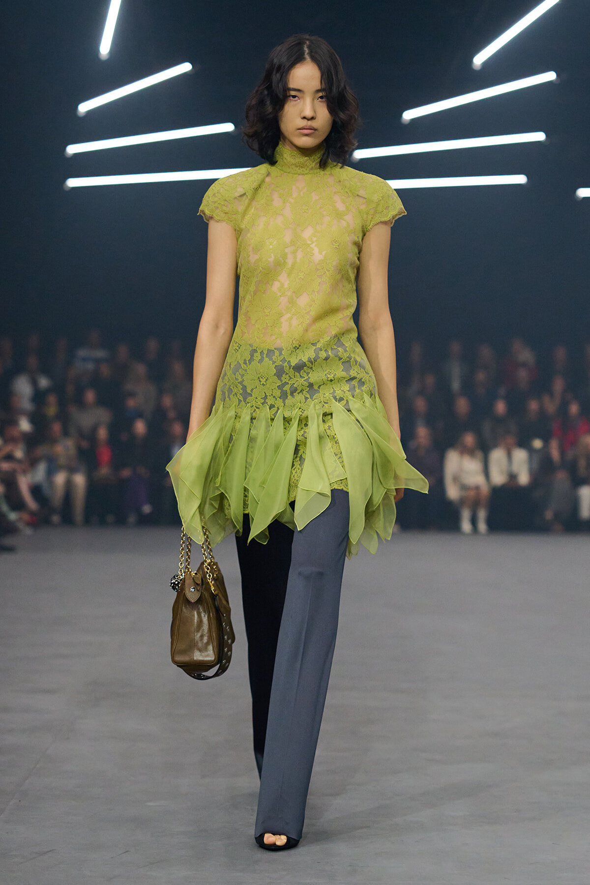 Model walking runway in lime lace high-neck top over sheer skirted hem, blue-gray wide-leg pants, brown handbag with chain.
