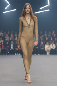Model walking the runway in a sheer nude-toned knit bodysuit with a deep V-neck, paired with pale pink heels; audience blurred in the background.