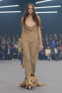 Model walks a beige, draped gown with silver chain embellishments down a runway.