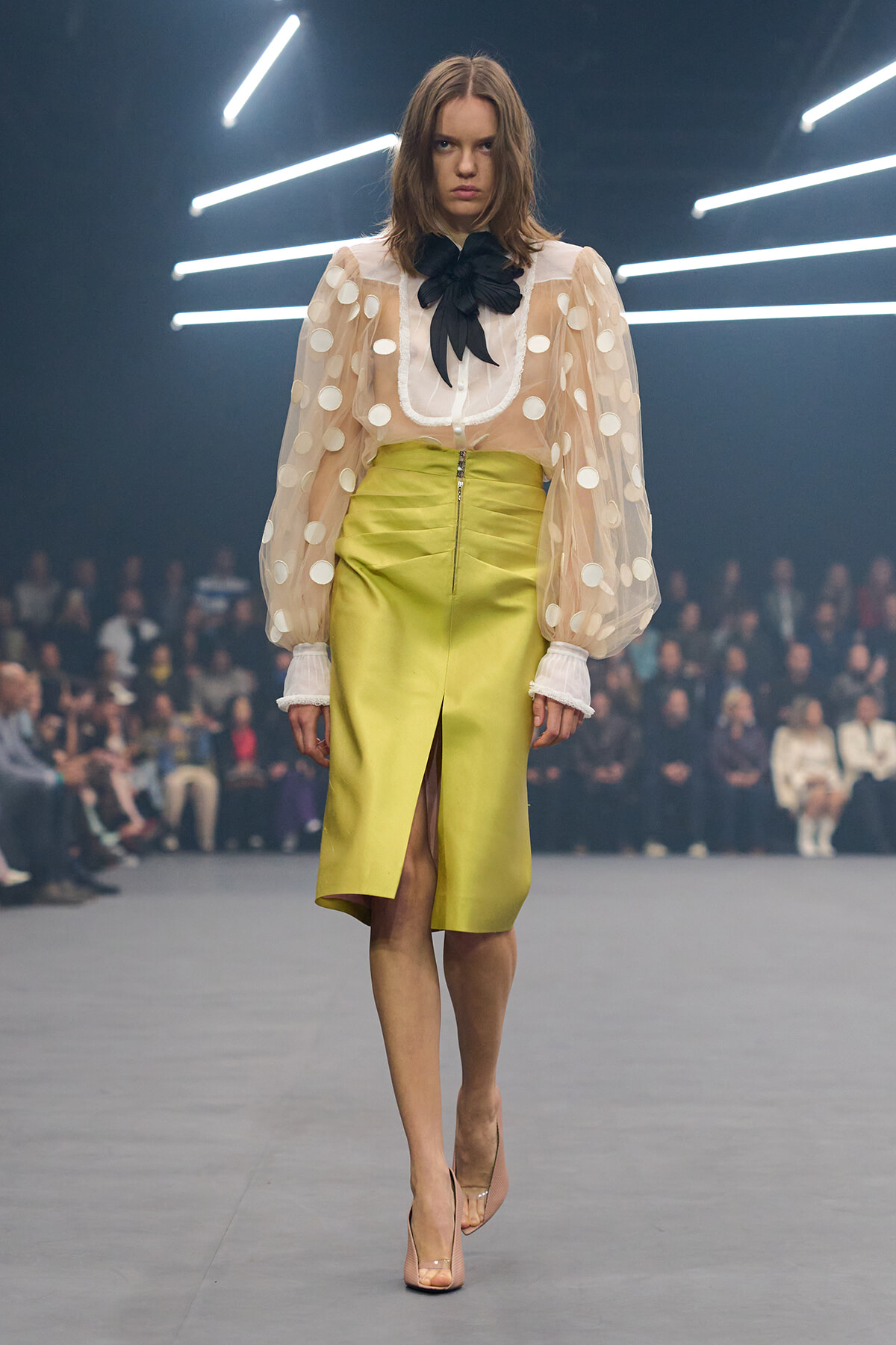 Runway model walking the catwalk in a sheer polka-dot blouse with a large black bow and a lime-green midi skirt, audience in background.
