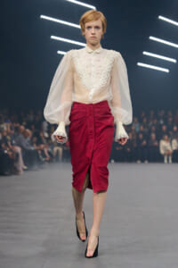 Model walks the runway in a sheer cream blouse with lace bib detailing and voluminous sleeves, paired with a red velvet midi skirt and black heels.