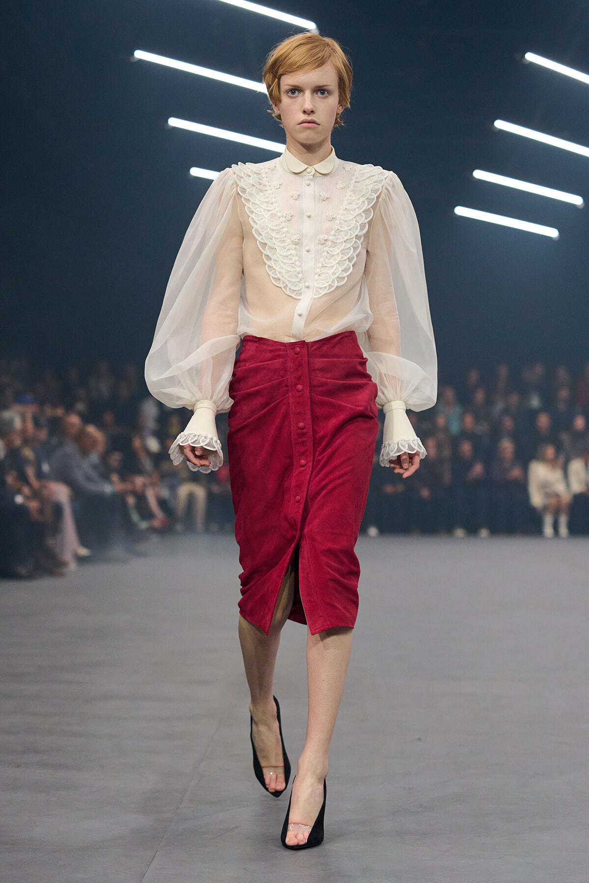 Model walks the runway in a sheer cream blouse with lace bib detailing and voluminous sleeves, paired with a red velvet midi skirt and black heels.