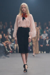 Model walking a fashion runway in a pink sheer blouse with voluminous sleeves, a black pencil skirt, and a large orange flower accessory at the neck.