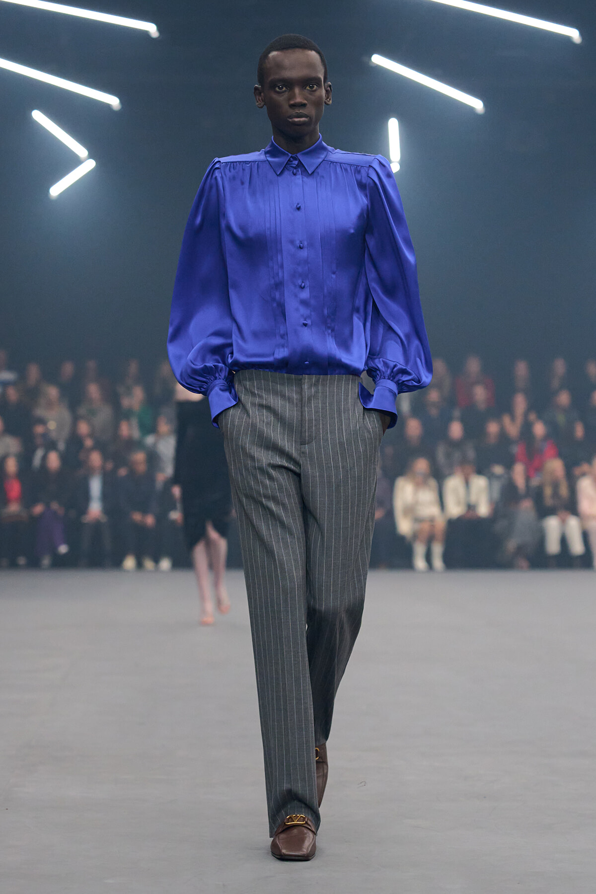 Model walking a runway in a cobalt blue satin blouse with puff sleeves and gray pinstripe trousers.