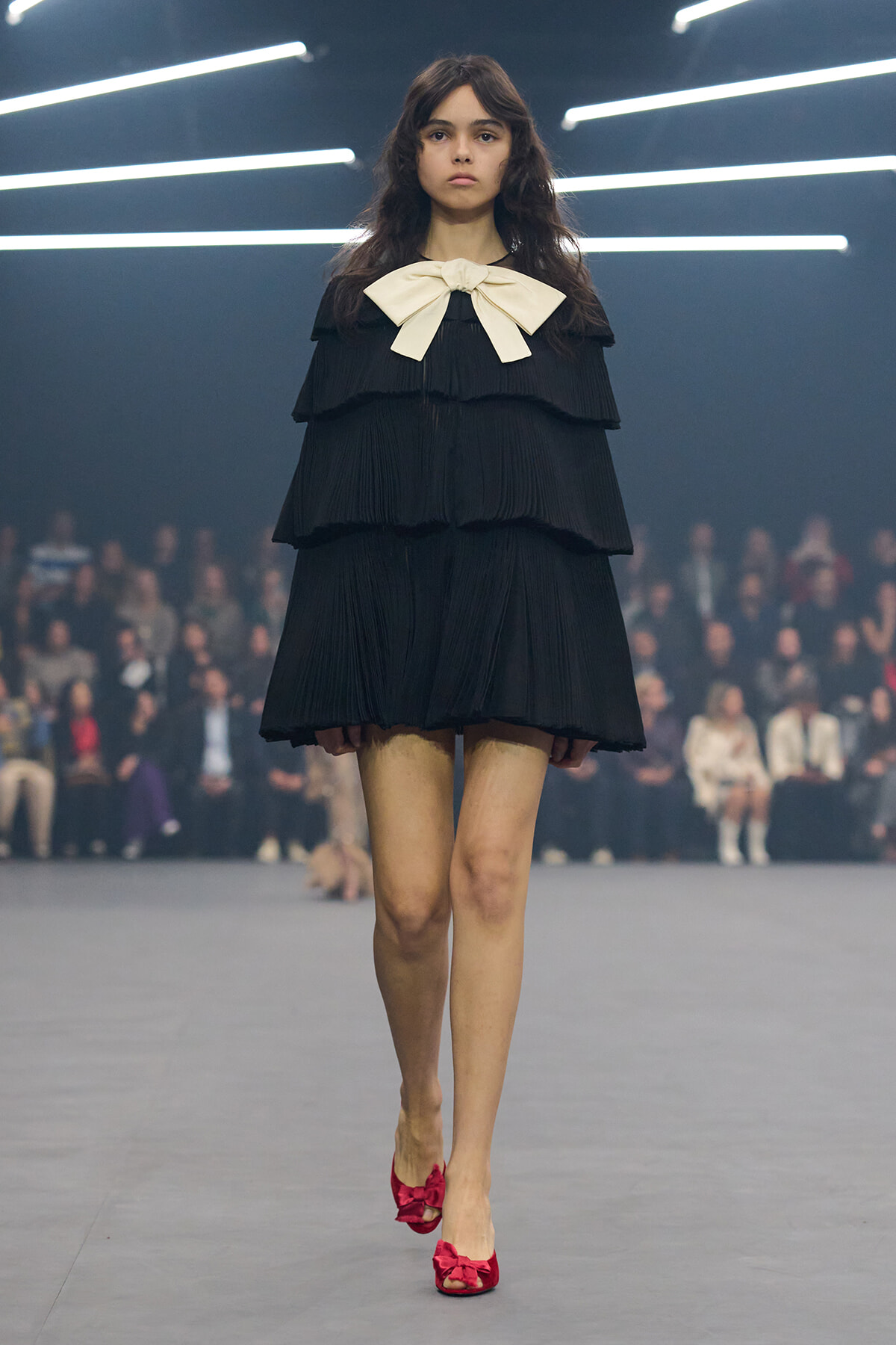 Model walking a fashion show in a black layered cape dress with a large ivory bow at the neckline and red bow-front heels.