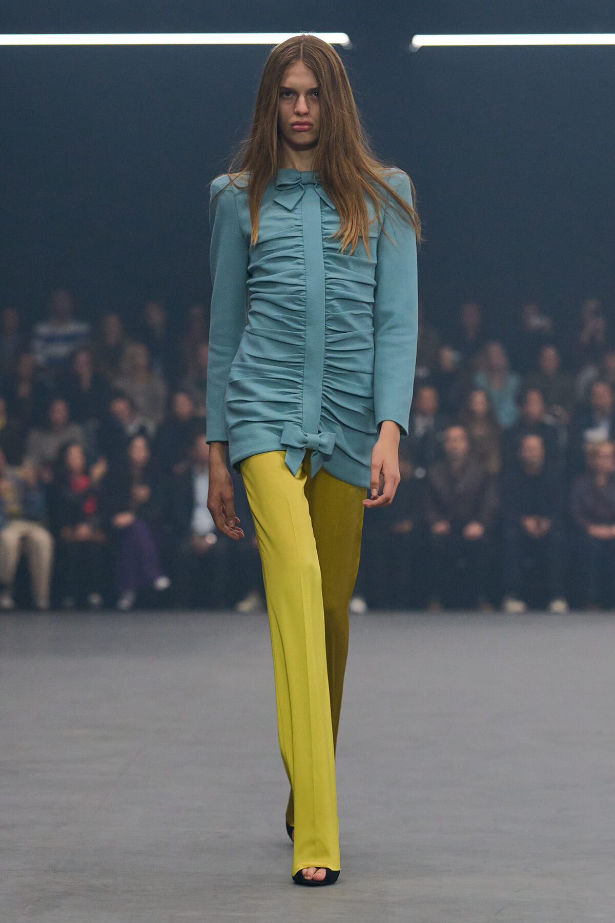 Model walking a fashion runway in a light blue ruched blouse with a bow and bright yellow wide-leg pants, audience blurred in the background.
