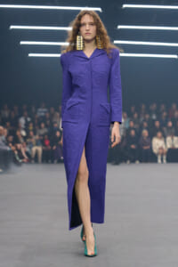 Female model walking a runway in a purple embossed coat with a front slit, large gold earrings, and teal heels.