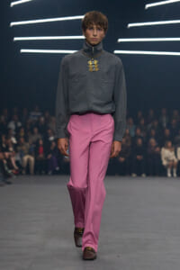 Male model on a runway wearing a gray shirt, pink trousers, and a bold gold pendant necklace, with an audience in the background.