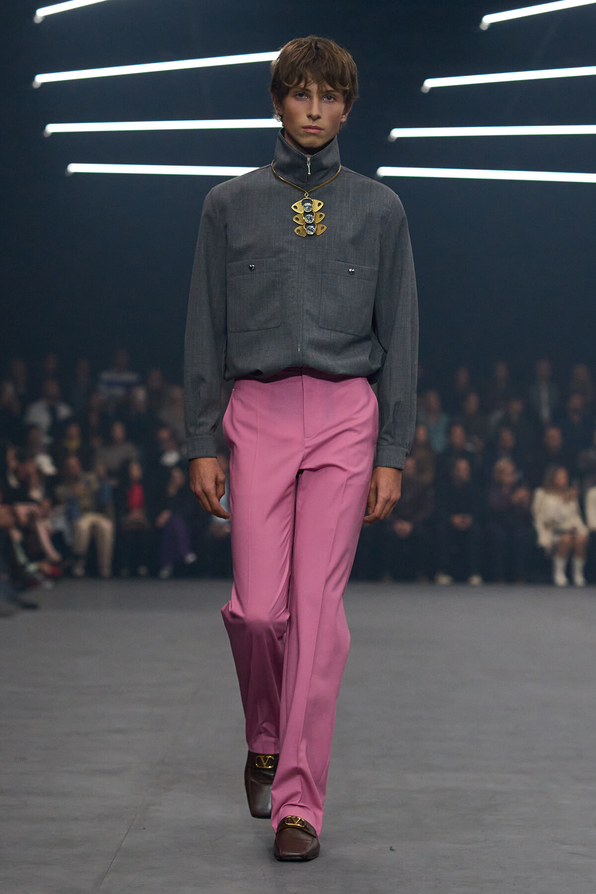 Male model on a runway wearing a gray shirt, pink trousers, and a bold gold pendant necklace, with an audience in the background.