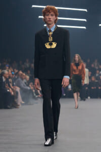 Male model walks a runway in a black double-breasted suit with a large gold statement necklace; audience blurred in background.