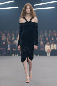 Model walking down a runway in a black velvet off-shoulder dress with front ruching and a center slit, audience blurred in the background.