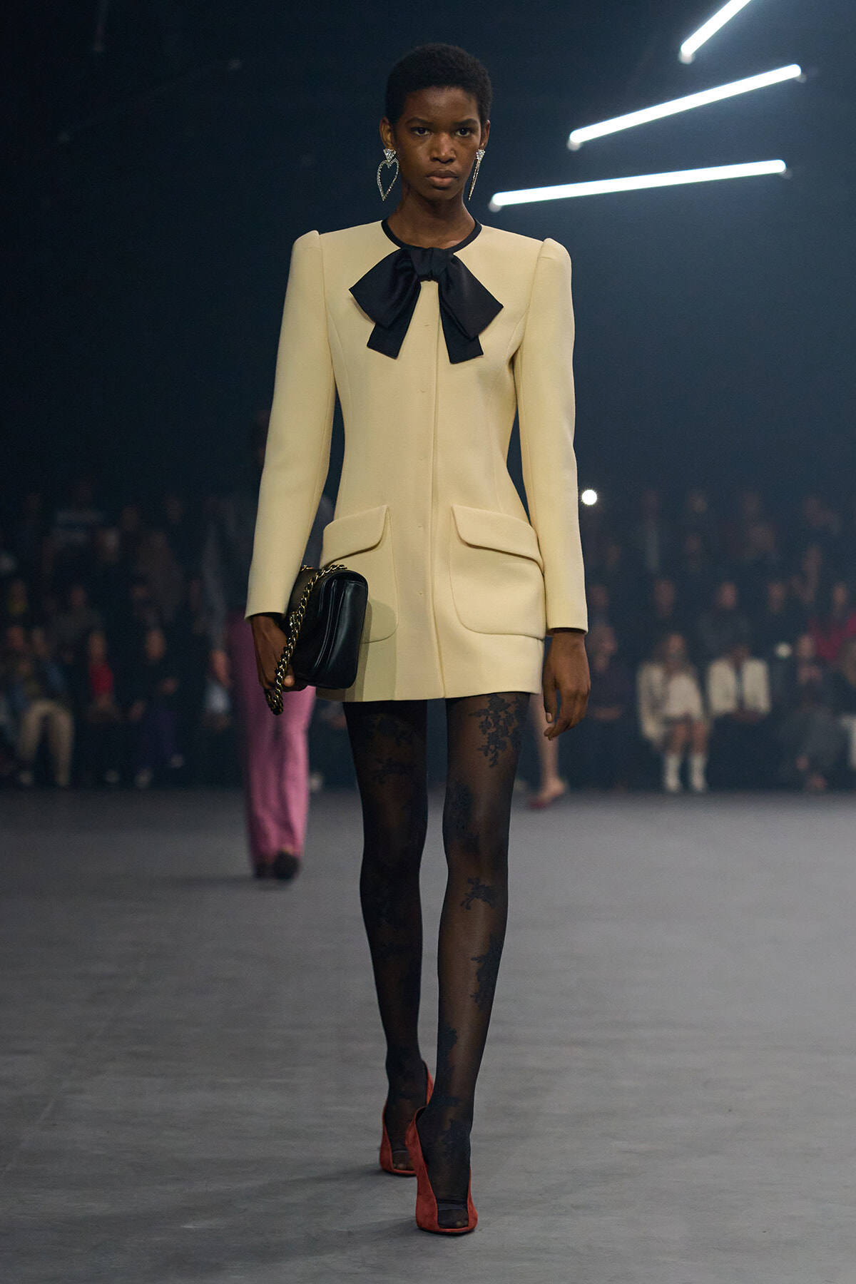 Model on a fashion runway wearing a cream tailored blazer dress with a large black bow at the neck, black patterned tights, and red heels, carrying a black clutch with a chain strap.