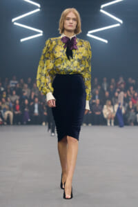 Model walking the runway in a yellow floral blouse with a purple bow, paired with a navy velvet pencil skirt and black heels.