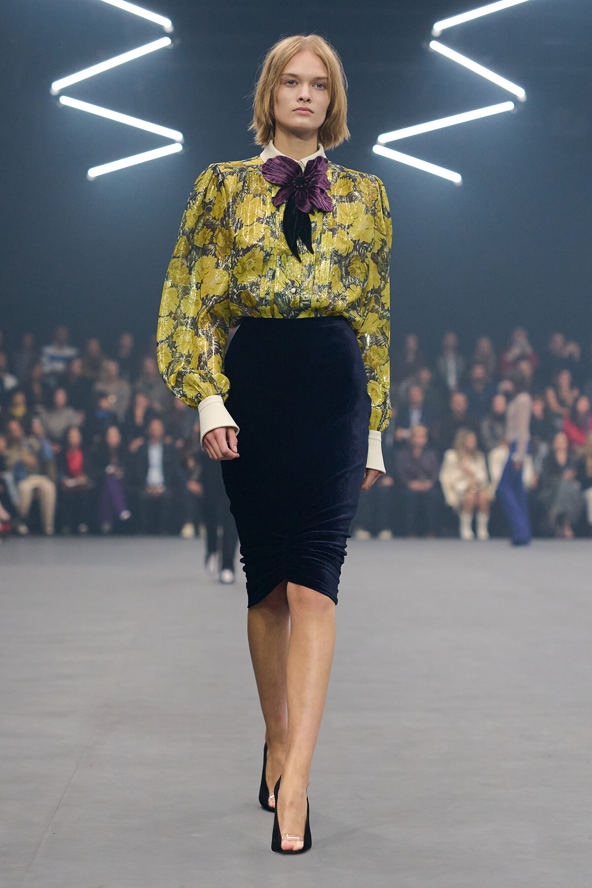 Model walking the runway in a yellow floral blouse with a purple bow, paired with a navy velvet pencil skirt and black heels.