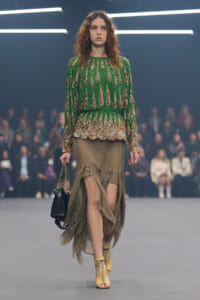 Model walks a fashion runway in a green sequined blouse with a gold scalloped hem and a taupe draped skirt, carrying a small black purse with a chain strap.