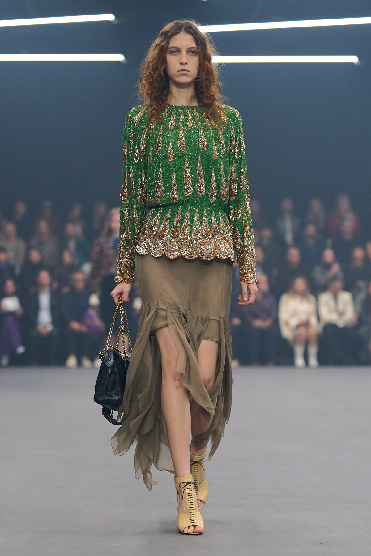 Model walks a fashion runway in a green sequined blouse with a gold scalloped hem and a taupe draped skirt, carrying a small black purse with a chain strap.