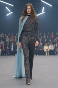 Model walks the runway in a glittery silver sequin jumpsuit with a pale blue cape draped over one shoulder.