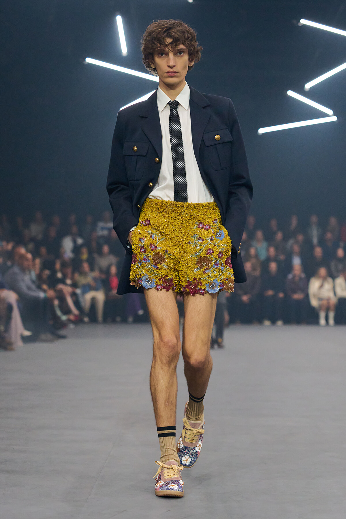 Male runway model in a navy blazer with gold buttons, white shirt, and a bright yellow floral-embroidered mini skirt.