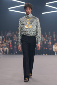 Male model on a runway wearing a glittering silver high-neck blouse, black trousers, and a large gold pendant necklace.