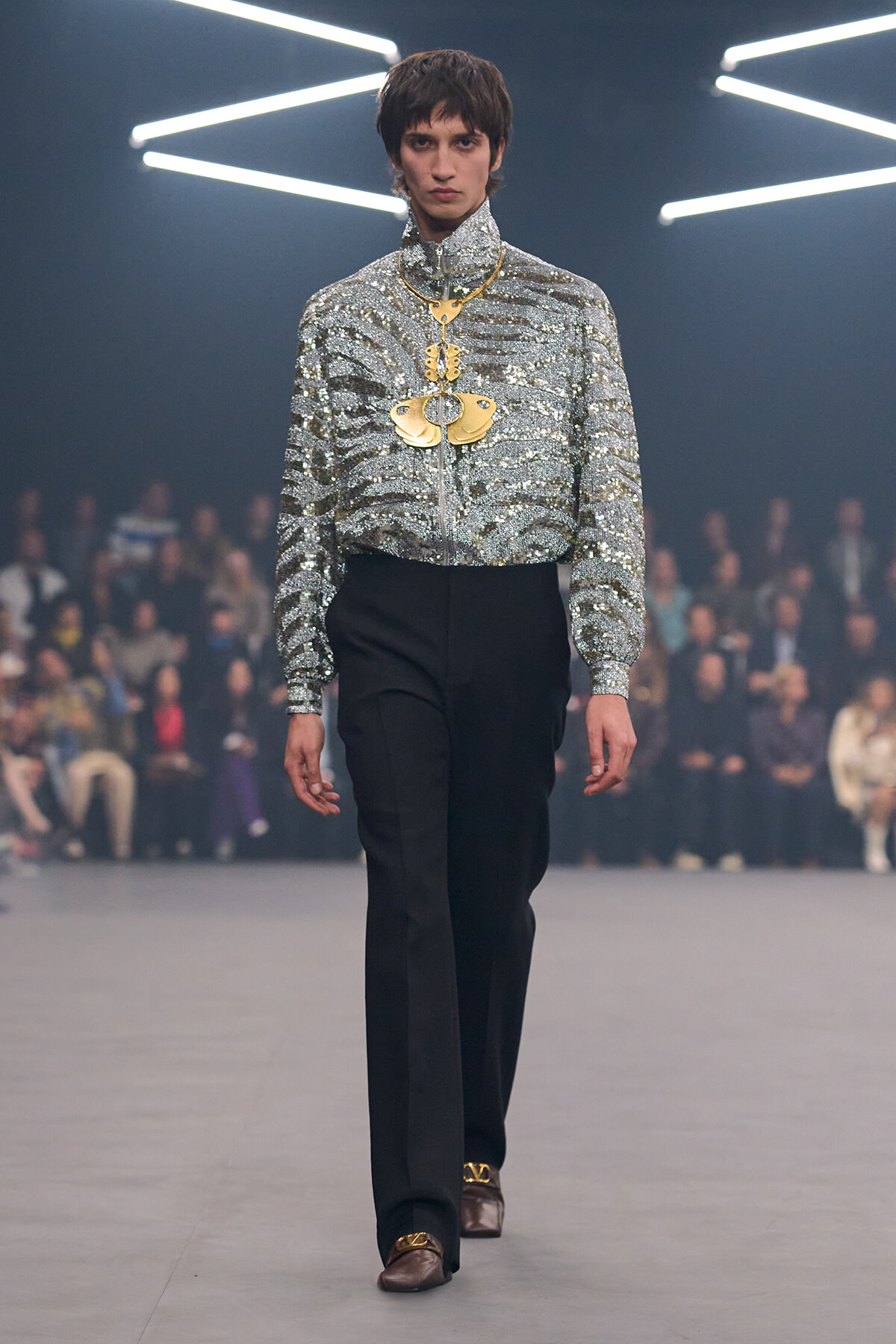 Male model on a runway wearing a glittering silver high-neck blouse, black trousers, and a large gold pendant necklace.