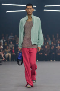 Model walks a runway in a mint bomber jacket, glittery pink top, pink wide-leg pants, blue purse, and embroidered shoes.