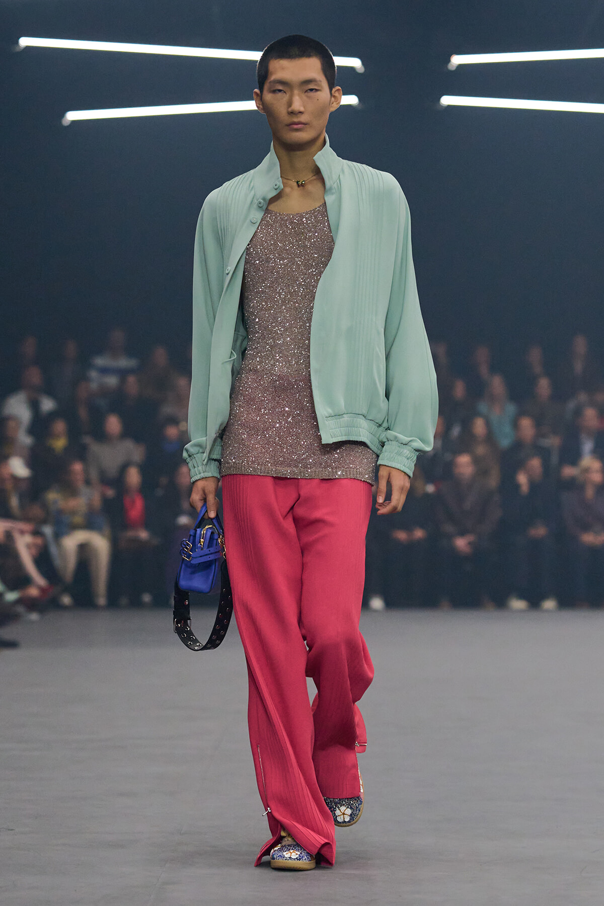Model walks a runway in a mint bomber jacket, glittery pink top, pink wide-leg pants, blue purse, and embroidered shoes.