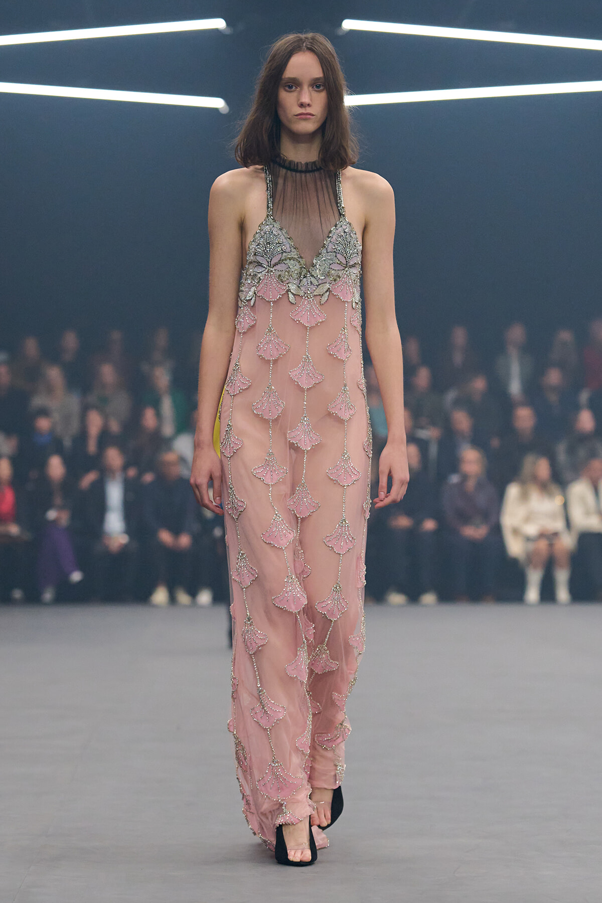 Model walking a pink, bead-embellished gown with a sheer neckline on a runway.