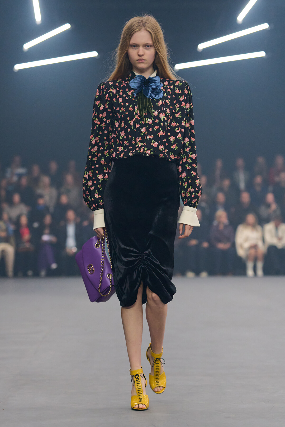 Model walking a fashion runway in a navy floral blouse, black velvet midi skirt, yellow lace-up sandals, and a purple handbag.