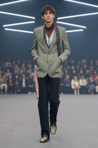 Male model walking a runway in a light gray blazer, layered colorful top, and patterned scarf with hands in pockets.