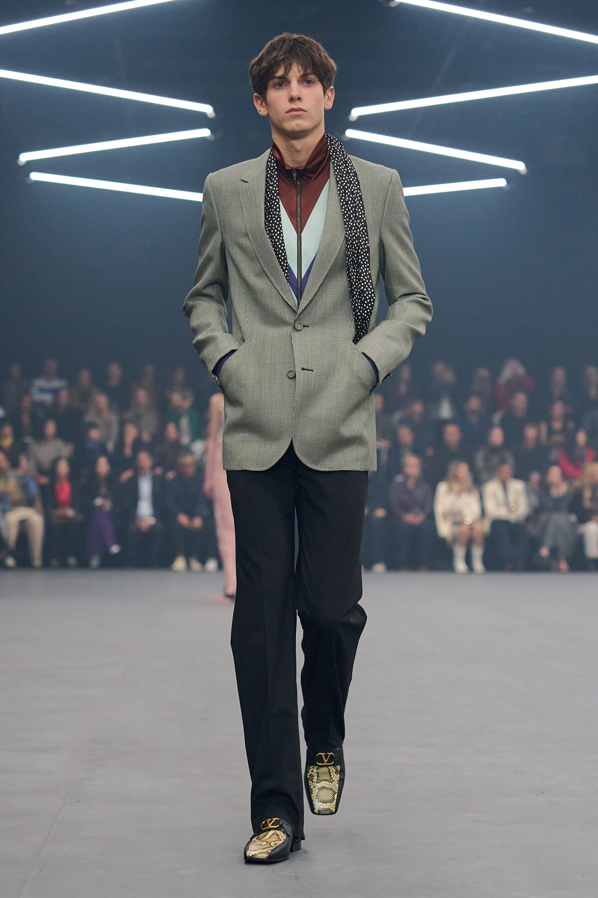 Male model walking a runway in a light gray blazer, layered colorful top, and patterned scarf with hands in pockets.