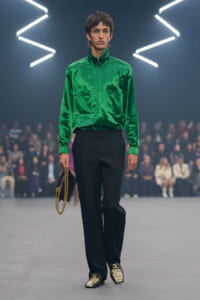 Male model on a fashion runway wearing an emerald satin shirt with chest pockets, black trousers, snakeskin loafers, and a gold chain handbag.