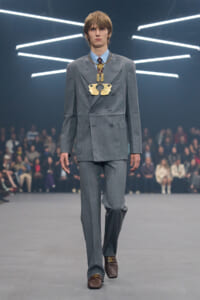 Male model walking a fashion runway in a light-gray double-breasted suit with an oversized gold necklace, brown loafers, against a dark backdrop with neon light bars.