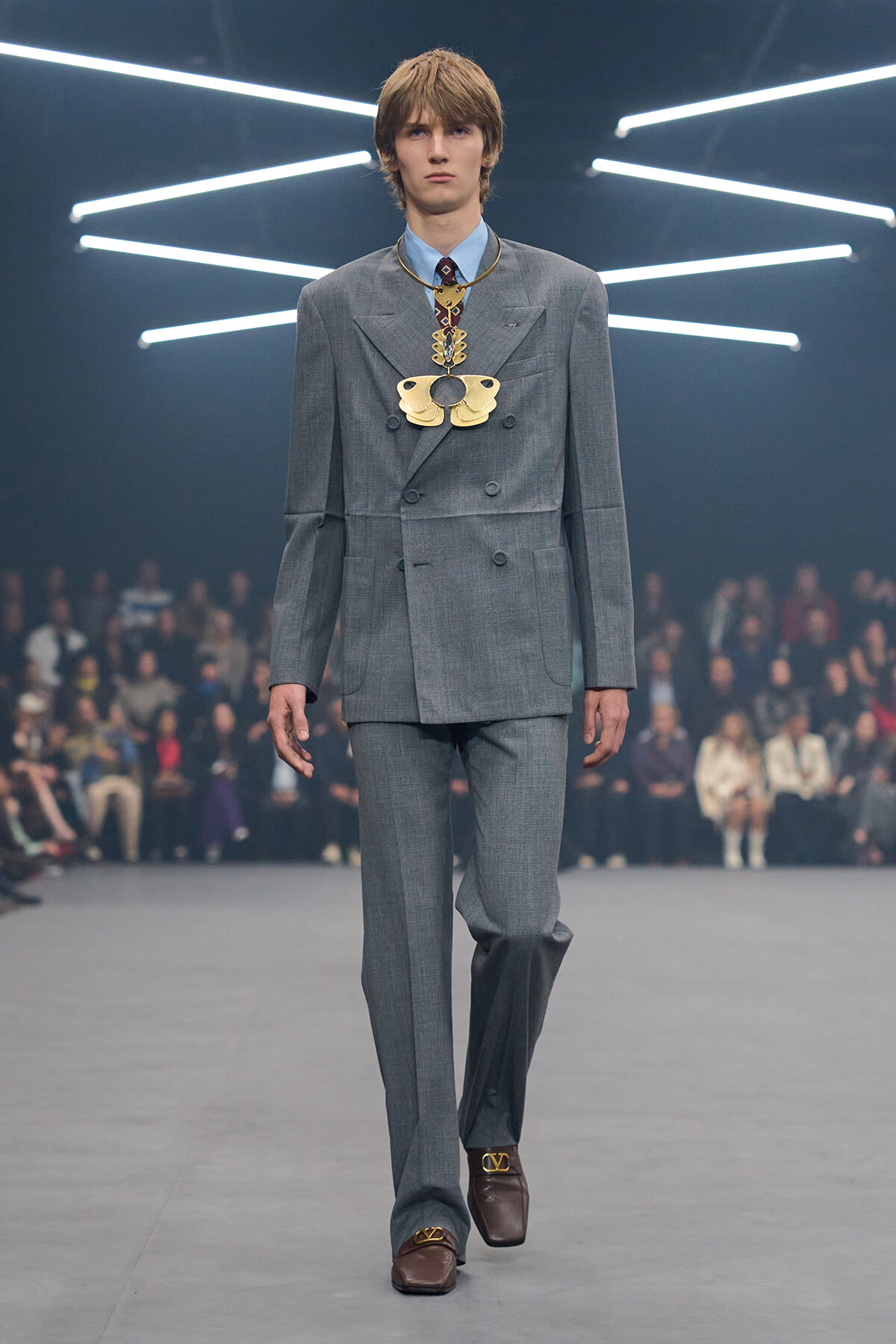 Male model walking a fashion runway in a light-gray double-breasted suit with an oversized gold necklace, brown loafers, against a dark backdrop with neon light bars.