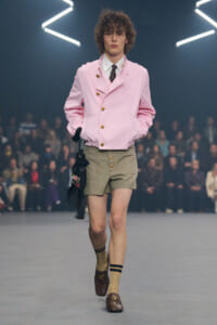 Male model walking a fashion runway in a pink double-breasted jacket with gold buttons, beige shorts, tan socks, and brown loafers, carrying a fringed bag.