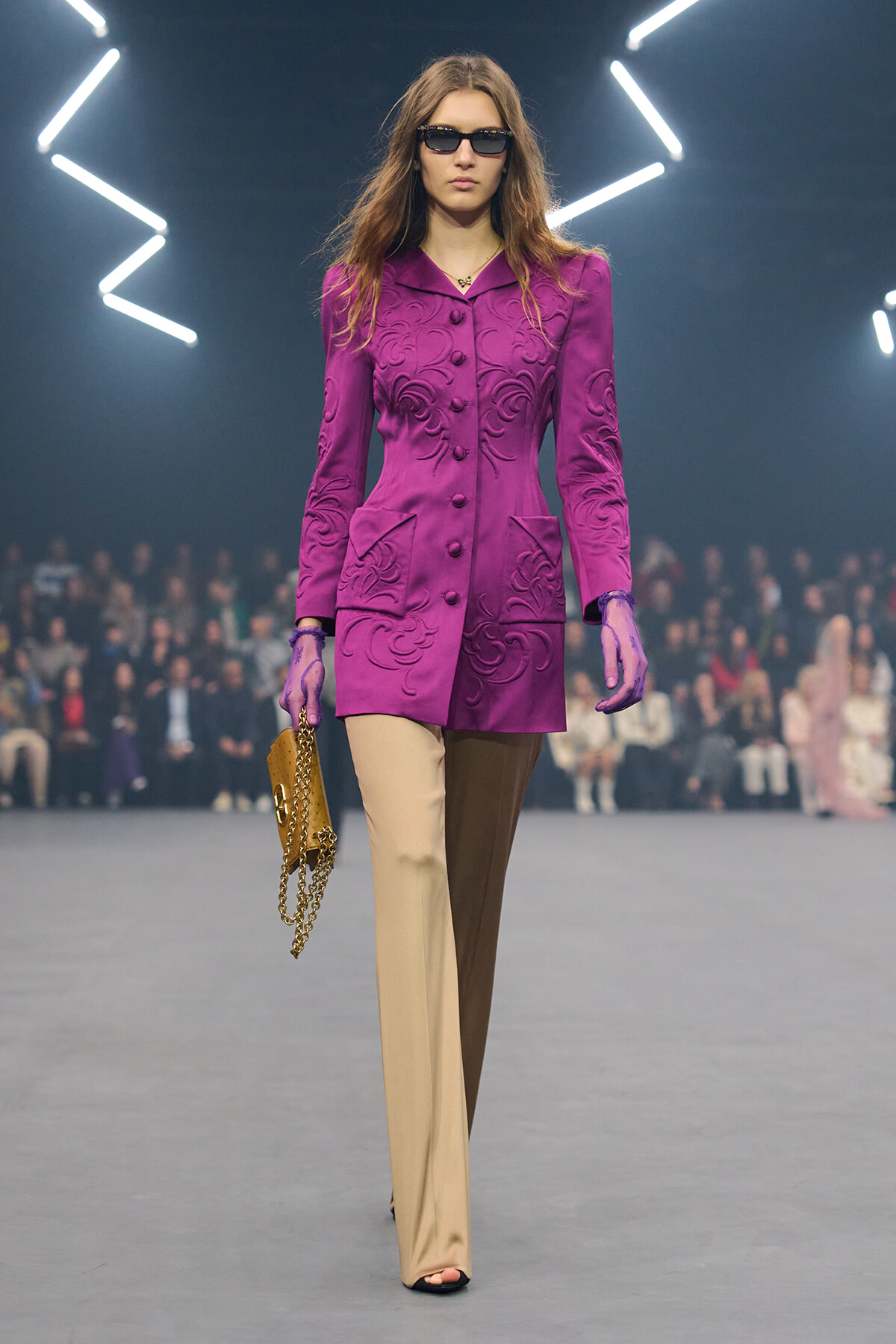 Model walking a runway in a vivid purple embroidered coat, two-tone beige and brown pants, black sunglasses, carrying a gold chain clutch.