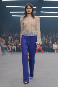 Model walking a fashion runway in a sheer striped blouse and royal blue wide-leg pants, audience blurred in the background,.