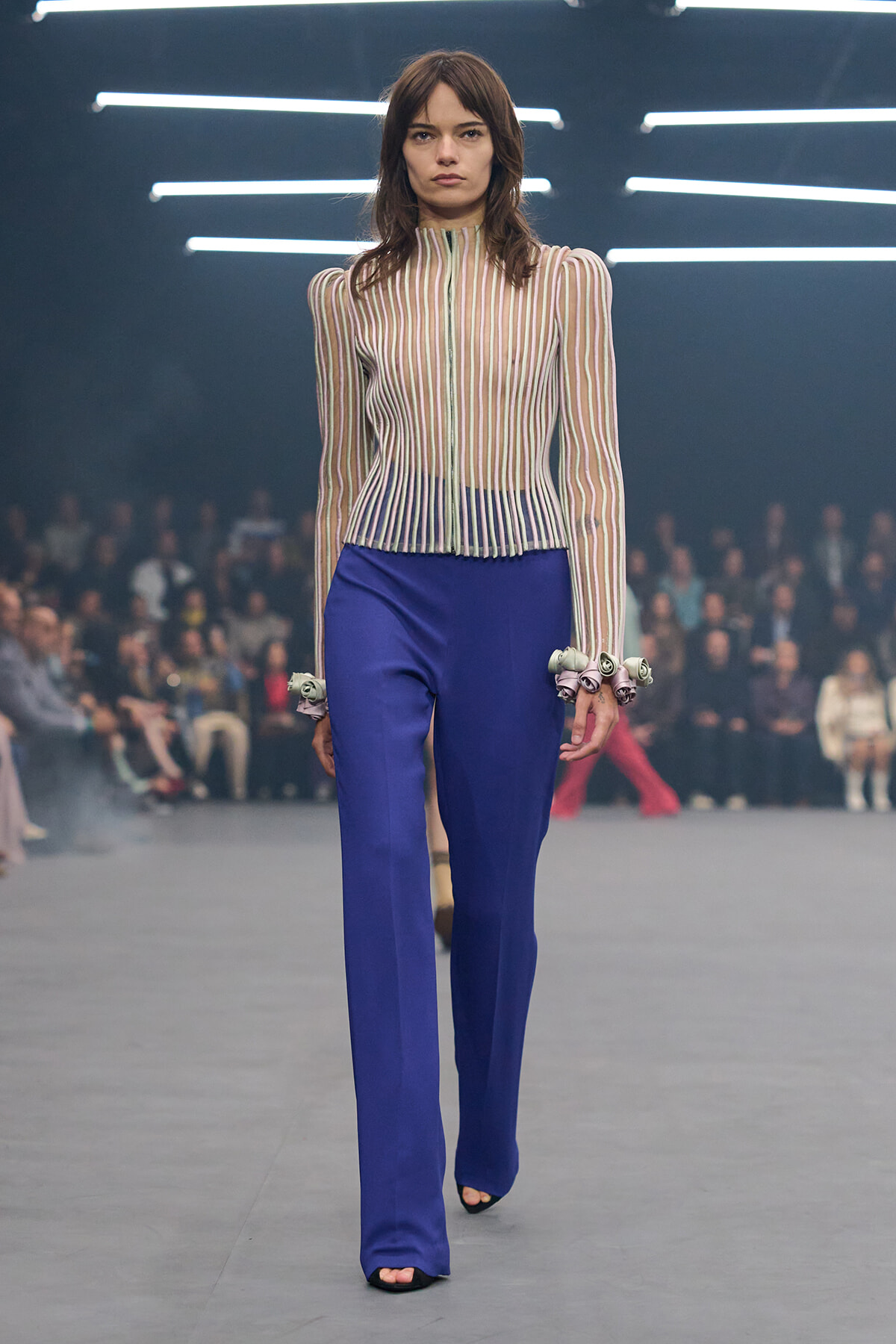 Model walking a fashion runway in a sheer striped blouse and royal blue wide-leg pants, audience blurred in the background,.