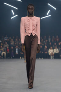 Model on a runway wearing a pink ruched blouse with a bow and brown wide-leg pants, audience in background.