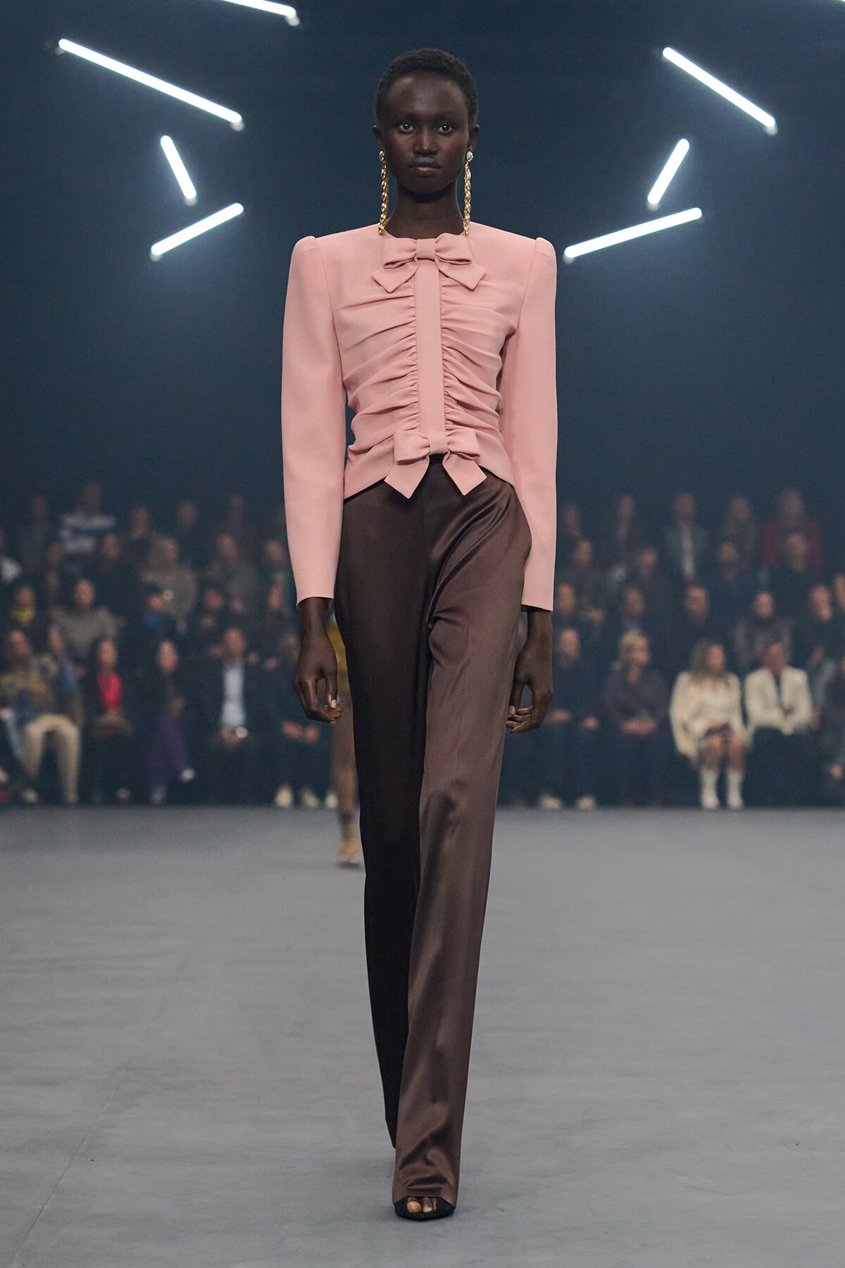 Model on a runway wearing a pink ruched blouse with a bow and brown wide-leg pants, audience in background.