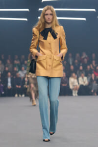 Model walking a fashion show in a tan coat with a black bow, light blue jeans, and a black handbag.