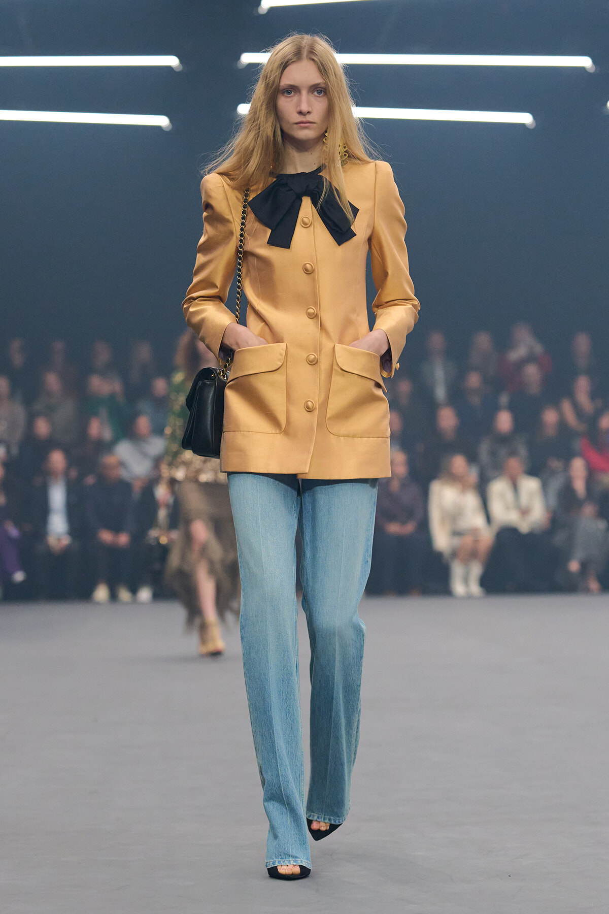 Model walking a fashion show in a tan coat with a black bow, light blue jeans, and a black handbag.