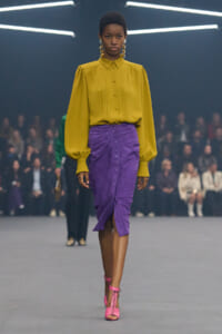 Model walking a fashion runway in a mustard yellow blouse with puff sleeves and a purple suede pencil skirt; pink strappy heels and statement earrings visible, audience in blurred background.